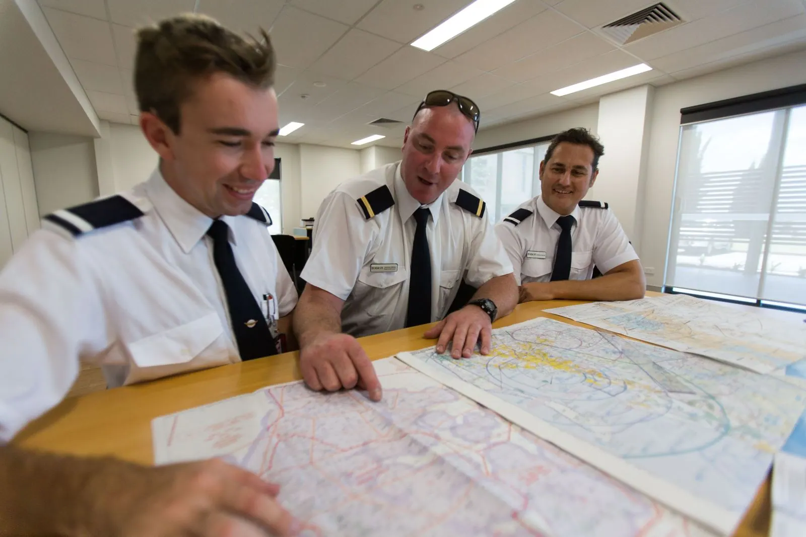 Three pilots in uniform planning a route over aeronautical charts spread across a classroom table